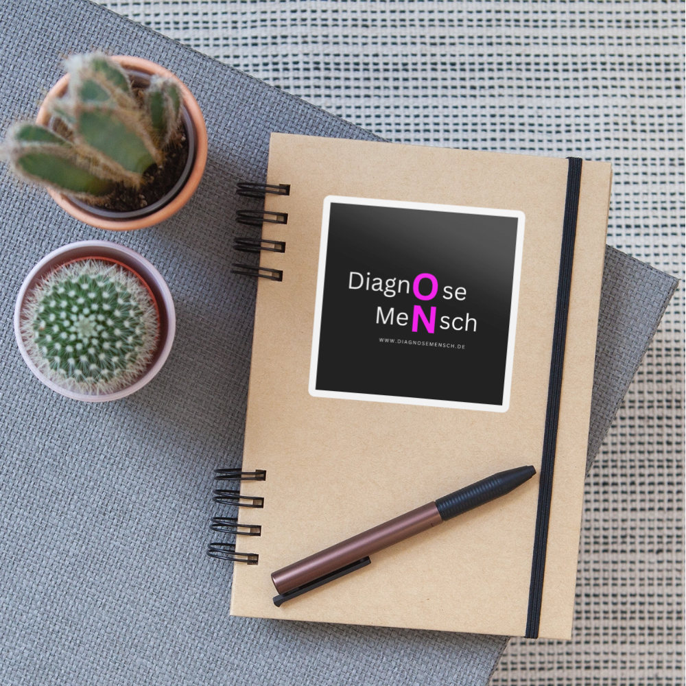 aufkleber, der auf einem notizbuch klebt. der aufkleber ist viereckig und hat einen schwarzen hintergrund. in weiß steht dort diagnose mensch und die buchstaben o und n sind in pink dargestellt für den begriff on.