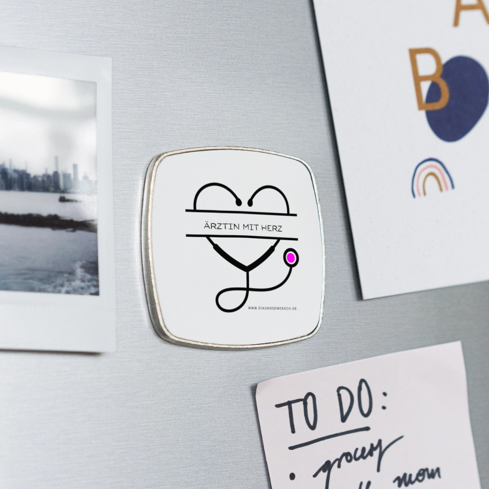 Ein Magnet mit einem Stethothoskop in Herzform und der Aufschrift Ärztin mit Herz ist an eine Kühlschranktür geheftet.